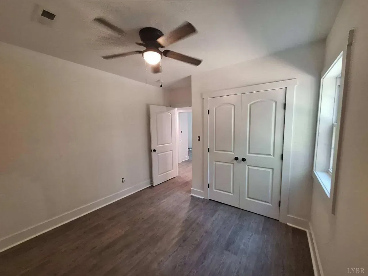 An empty bedroom with wood-look flooring, white walls, a ceiling fan, a window, and double doors.