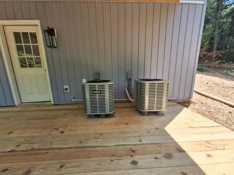 Two gray air conditioning units stand on a wooden deck next to a white door against a gray vertical-sided building.