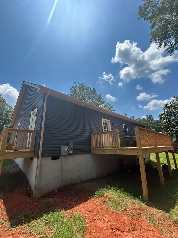 A house with dark grey siding and two wooden decks, situated on a grassy lot with red clay soil under a bright blue sky.