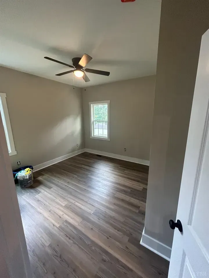 An empty bedroom with tan walls, wood-look flooring, a ceiling fan with a light, and a white window looking outside.