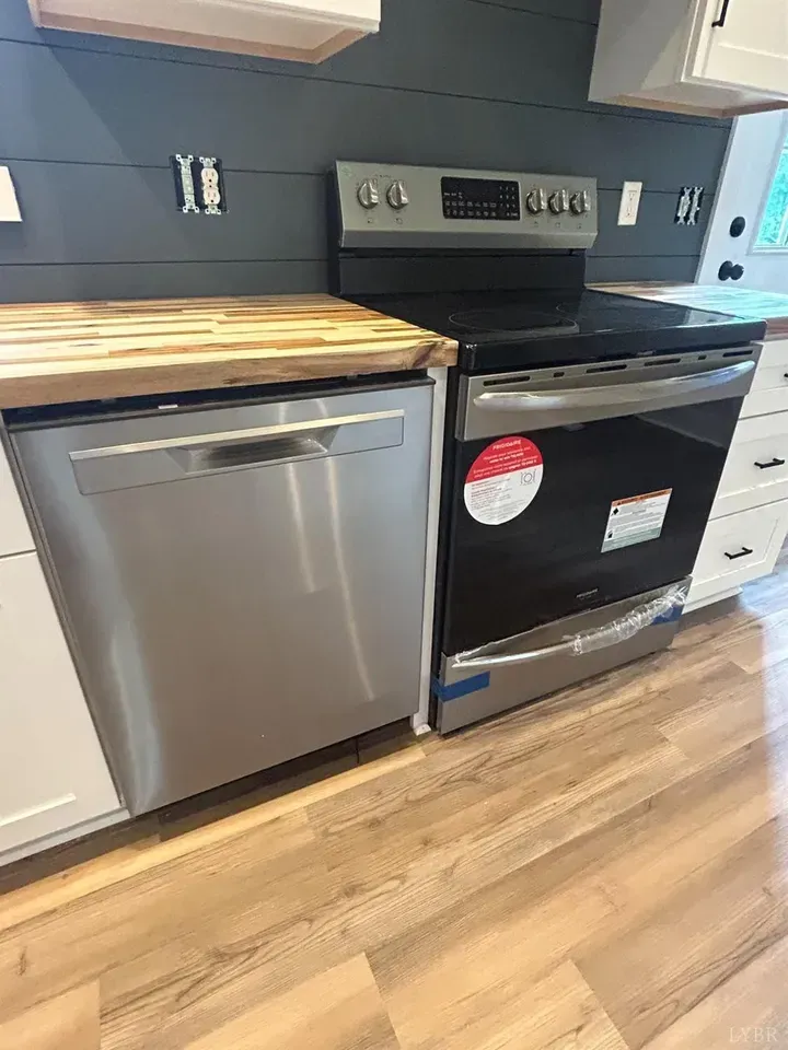 A stainless steel dishwasher and electric range sit side-by-side in a kitchen with white cabinets and dark shiplap walls.