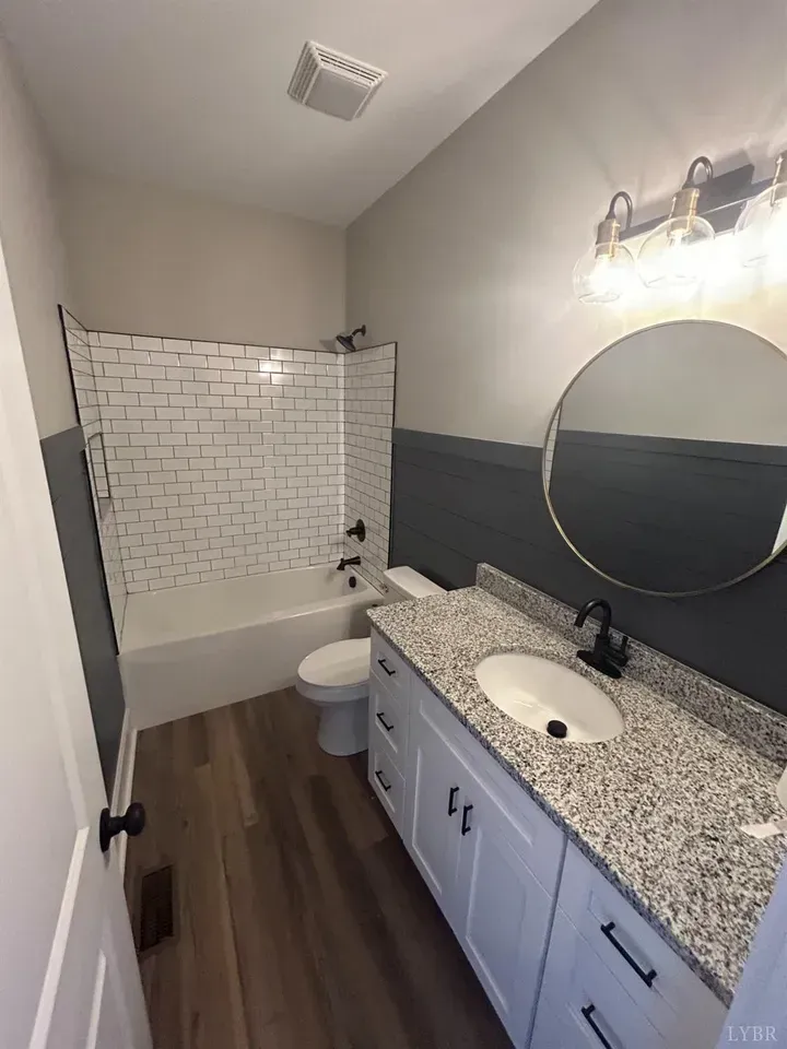 A modern bathroom featuring a white vanity with granite countertops, a round mirror, and a tub with white subway tiles.