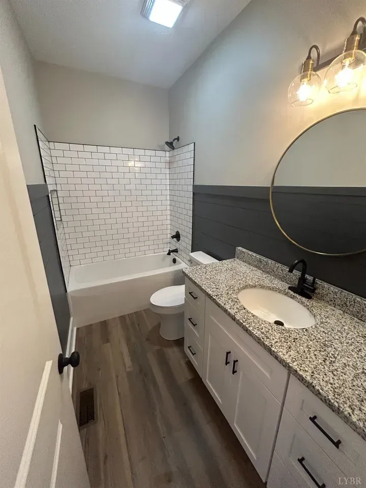 A modern bathroom featuring a white vanity with a granite countertop, a round mirror, and a white subway tile tub surround.