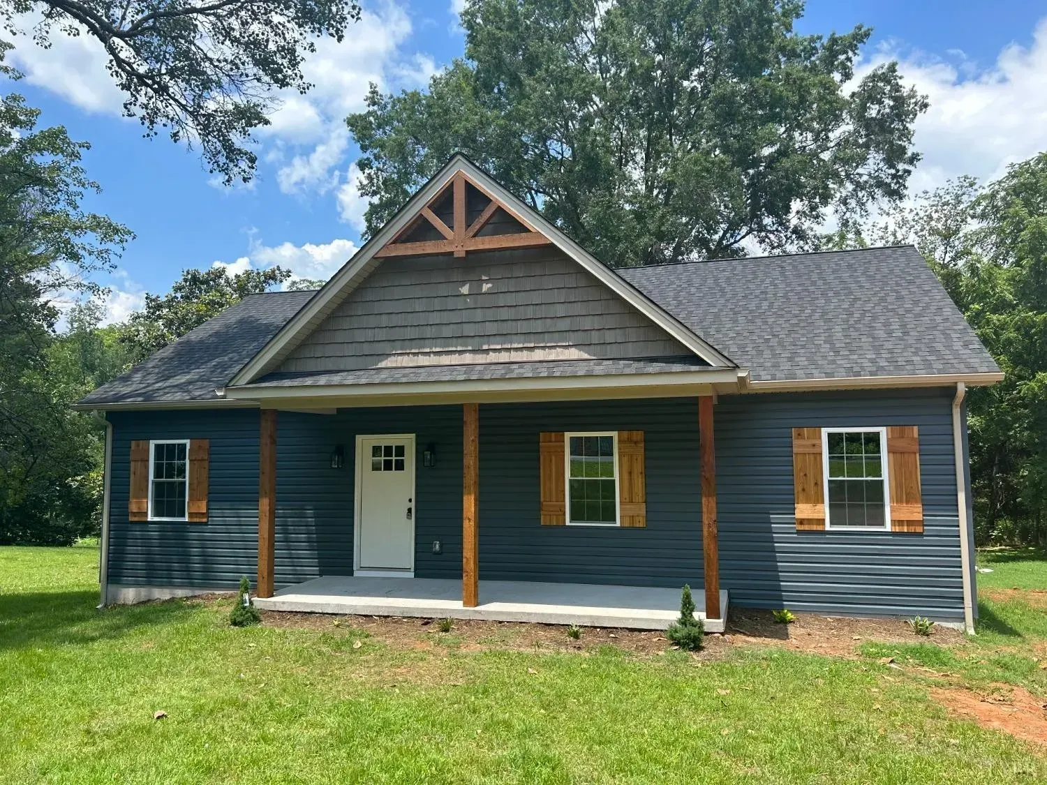 A single-story, dark blue house with a front porch, wood-tone shutters, and a triangular gable, set in a grassy yard.
