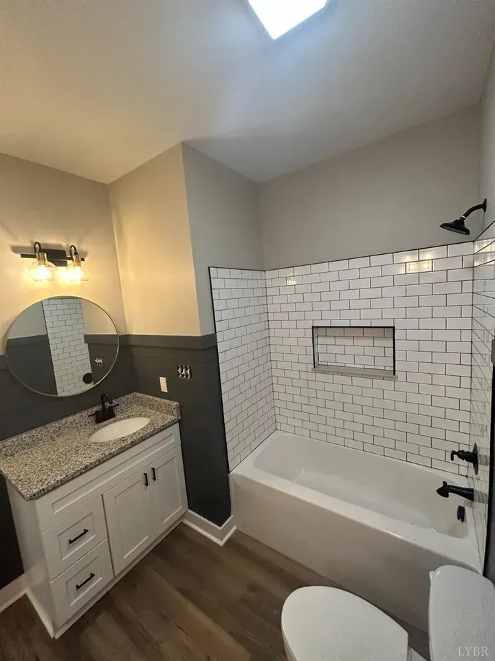A modern bathroom with a white vanity, speckled granite countertop, round mirror, and a white subway tile tub surround.