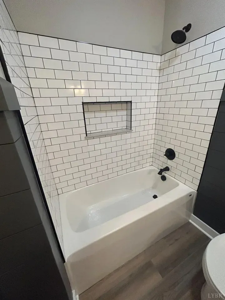 A bathroom features a white subway-tile shower enclosure with a recessed shelf, black fixtures, and a white bathtub.