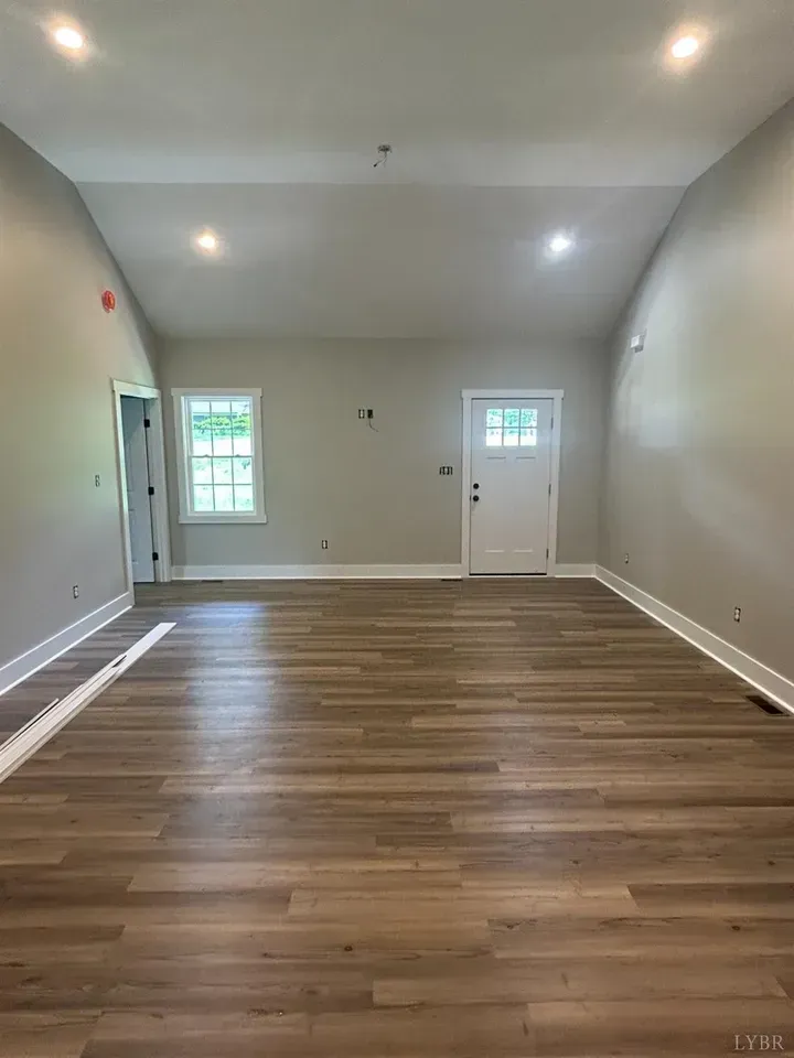 An empty room with light gray walls, a high vaulted ceiling, wood-look flooring, a window, and a white exterior door.