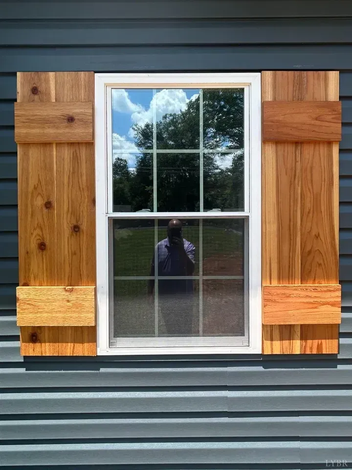 A white window with grid panes is flanked by rustic, natural wood shutters on dark blue horizontal siding.