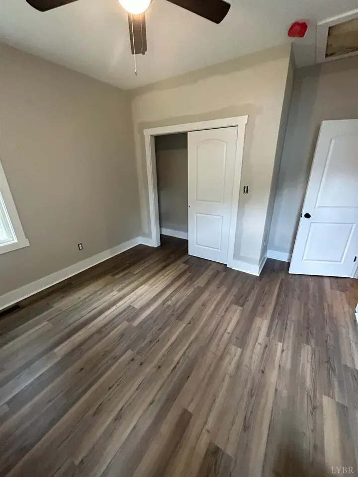 An empty room with light gray walls, brown wood-look flooring, a ceiling fan, a closet doorway, and a white interior door.