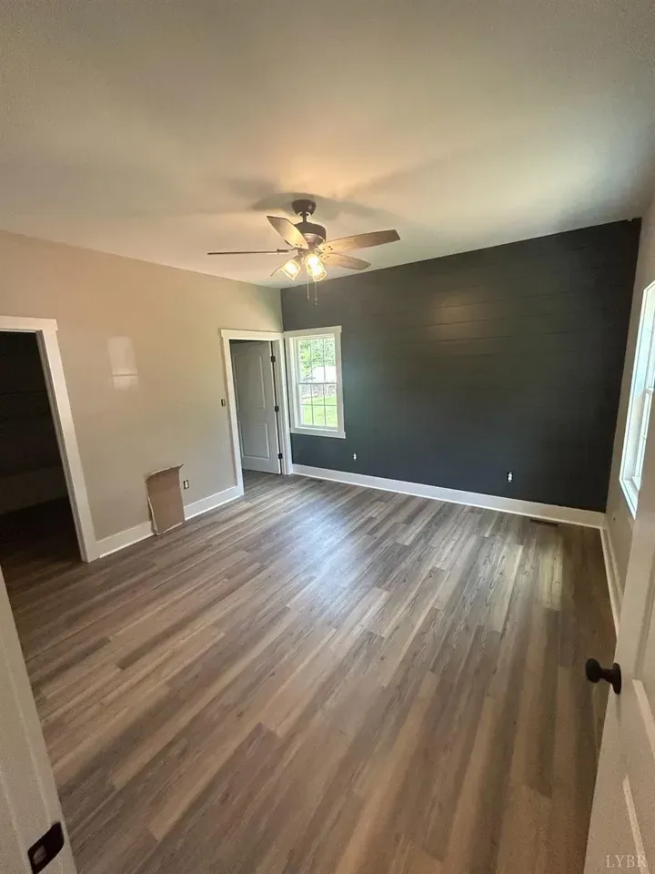 An empty room with wood-look flooring, light-colored walls, a dark accent wall, and a ceiling fan.