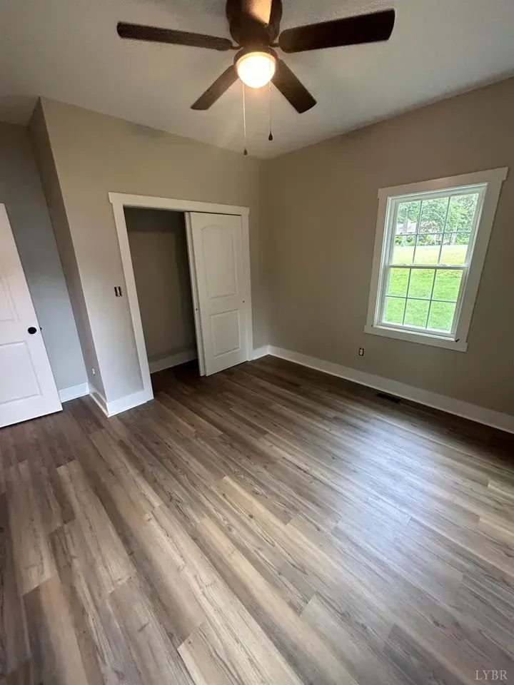 An empty bedroom with light gray walls, wood-look flooring, a ceiling fan, a closet, and a white-trimmed window.