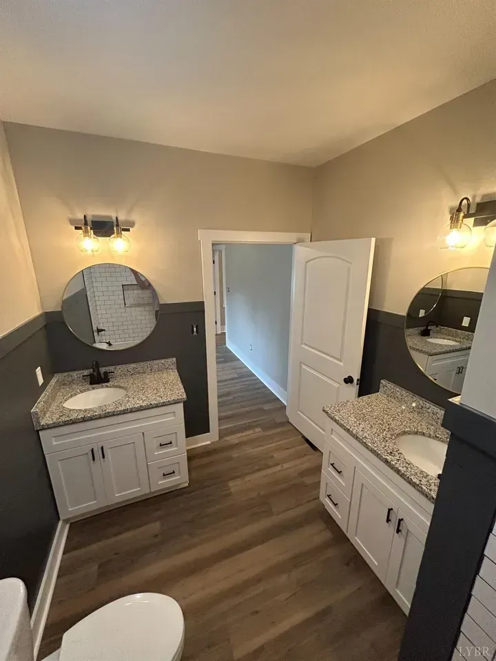 A bathroom with two white vanities, granite countertops, round mirrors, dark gray lower walls, and wood-look flooring.