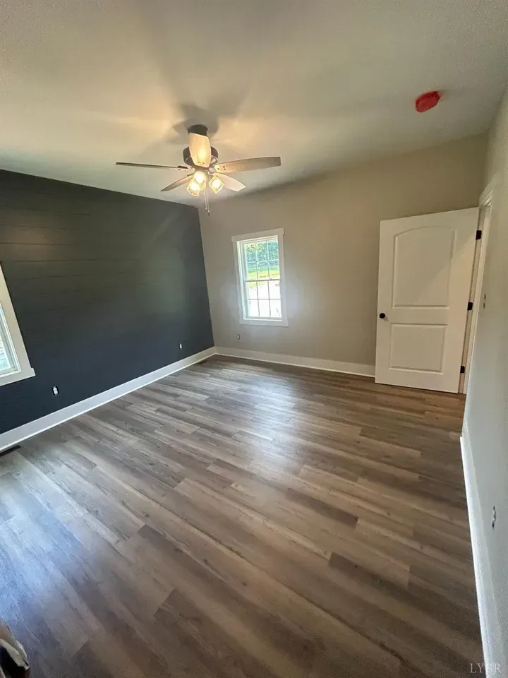 An empty room with light wood-look flooring, a dark charcoal accent wall, a ceiling fan, a window, and a white door.