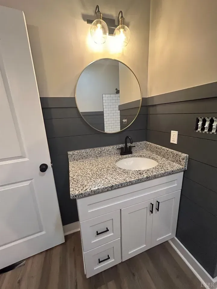 Modern bathroom vanity with a speckled countertop, white cabinetry, dark shiplap walls, a round mirror, and wall lighting.