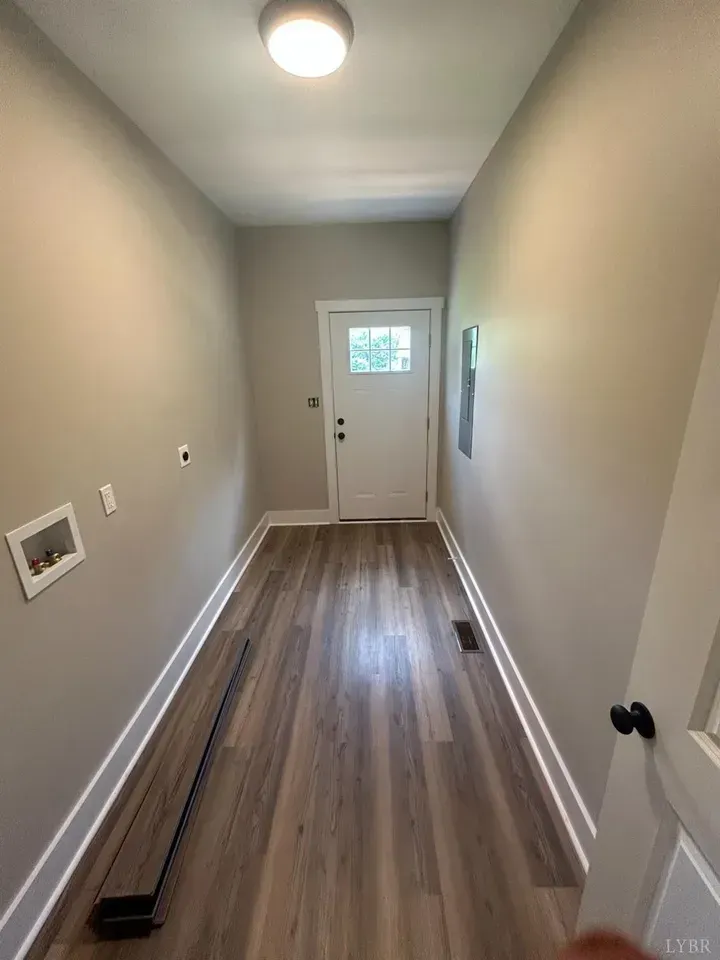 A narrow laundry room with light gray walls, wood-look flooring, a ceiling light, and a white exterior door at the end.