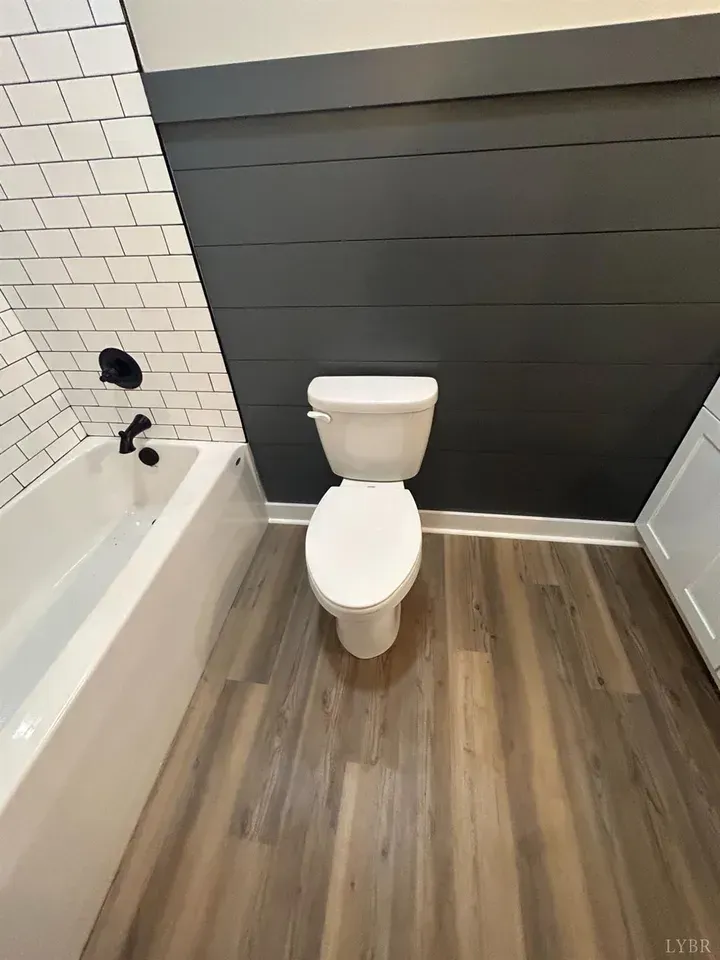 A bathroom with wood-look flooring, a white bathtub with black fixtures, and a toilet against a dark grey shiplap wall.