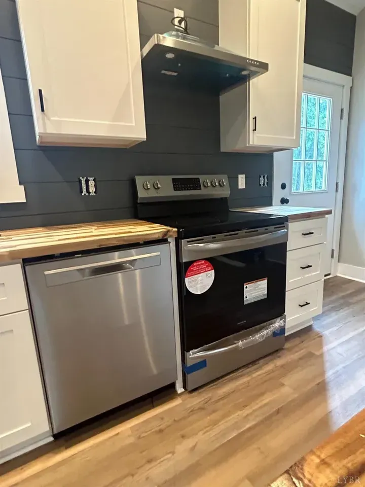 A stainless steel dishwasher and range stand beneath white cabinets and a range hood against a dark gray shiplap wall.