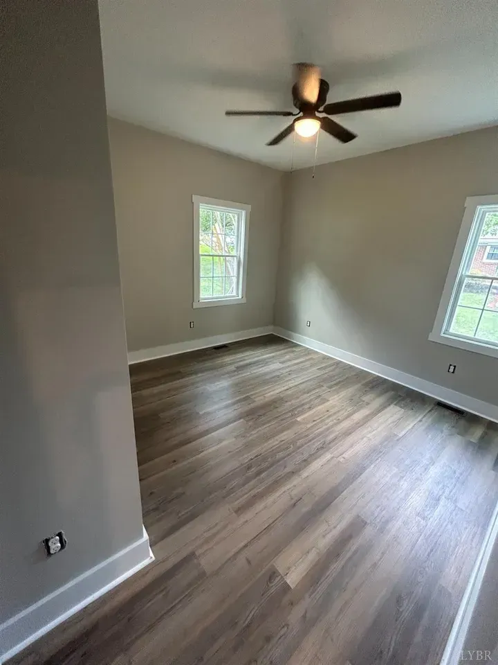 Empty room with neutral gray walls, wood-look flooring, two windows, and a ceiling fan.