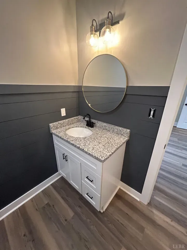 A modern bathroom vanity with a granite countertop, circular mirror, and dark grey shiplap walls over wood-look flooring.