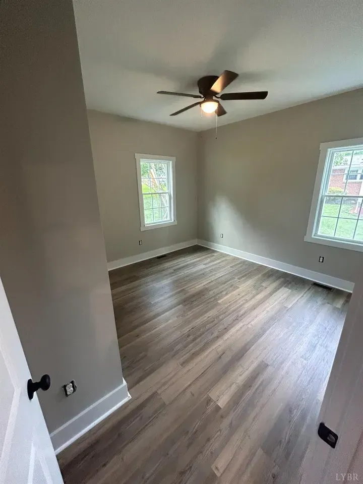An empty room with light gray walls, brown wood-look flooring, two windows, and a ceiling fan.