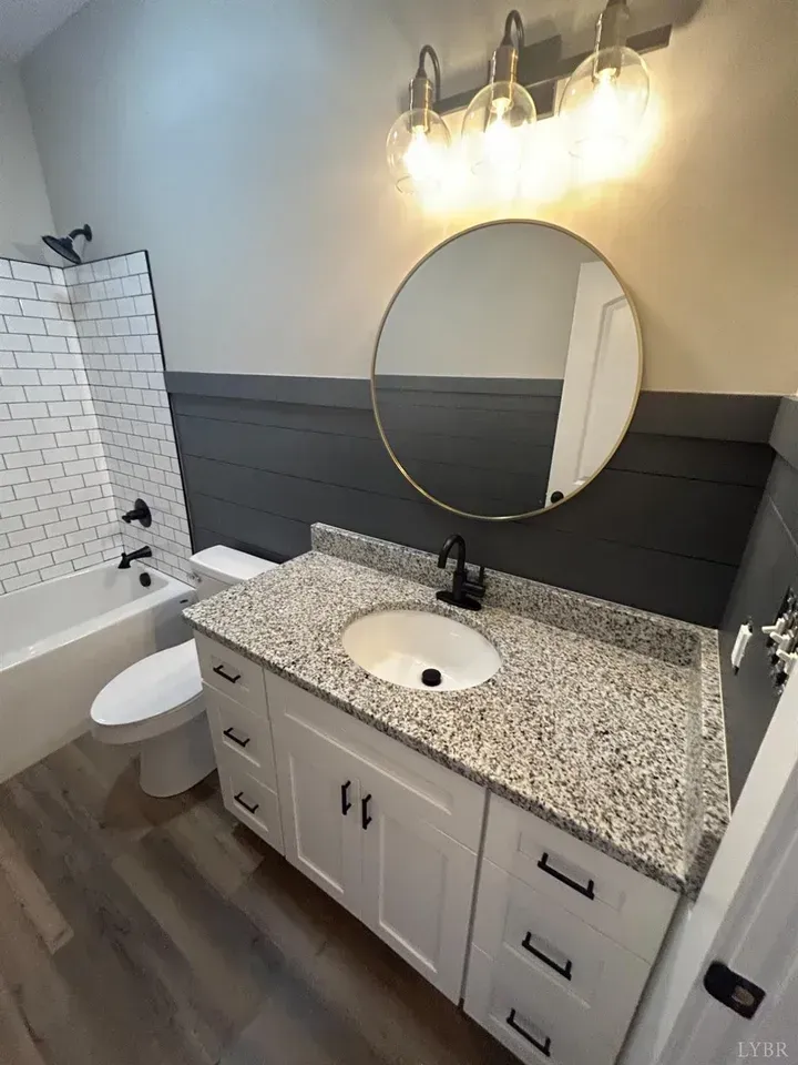 A modern bathroom features a white vanity with granite countertops, a round mirror, dark grey shiplap, and a tub-shower.