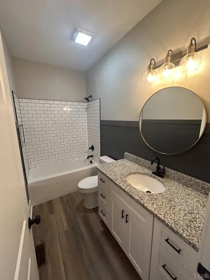 A modern bathroom featuring a white vanity with a granite countertop, circular mirror, and white subway-tiled bathtub.