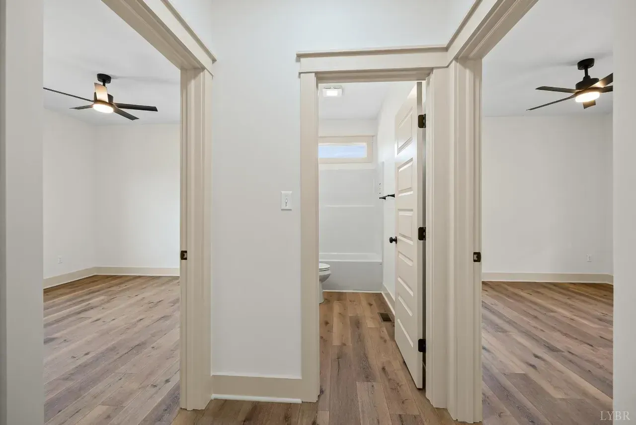 A bright hallway with light wood flooring leads to a central bathroom door, flanked by two doorways to empty bedrooms.