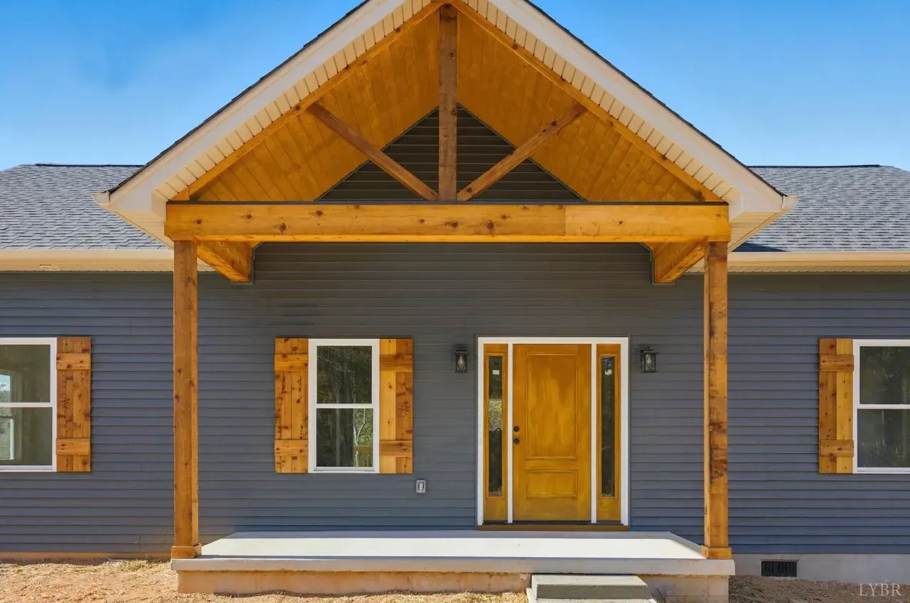 A front-facing view of a blue-sided house with a natural wood front door, rustic shutters, and a peaked wooden porch roof.