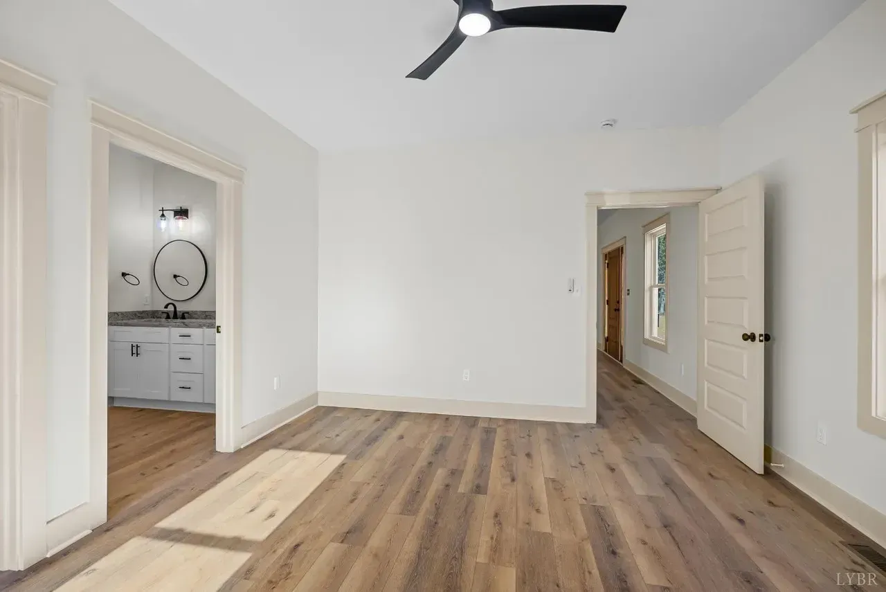 A bright, empty bedroom with white walls, wood-look flooring, a black ceiling fan, and doorways leading to a bathroom.