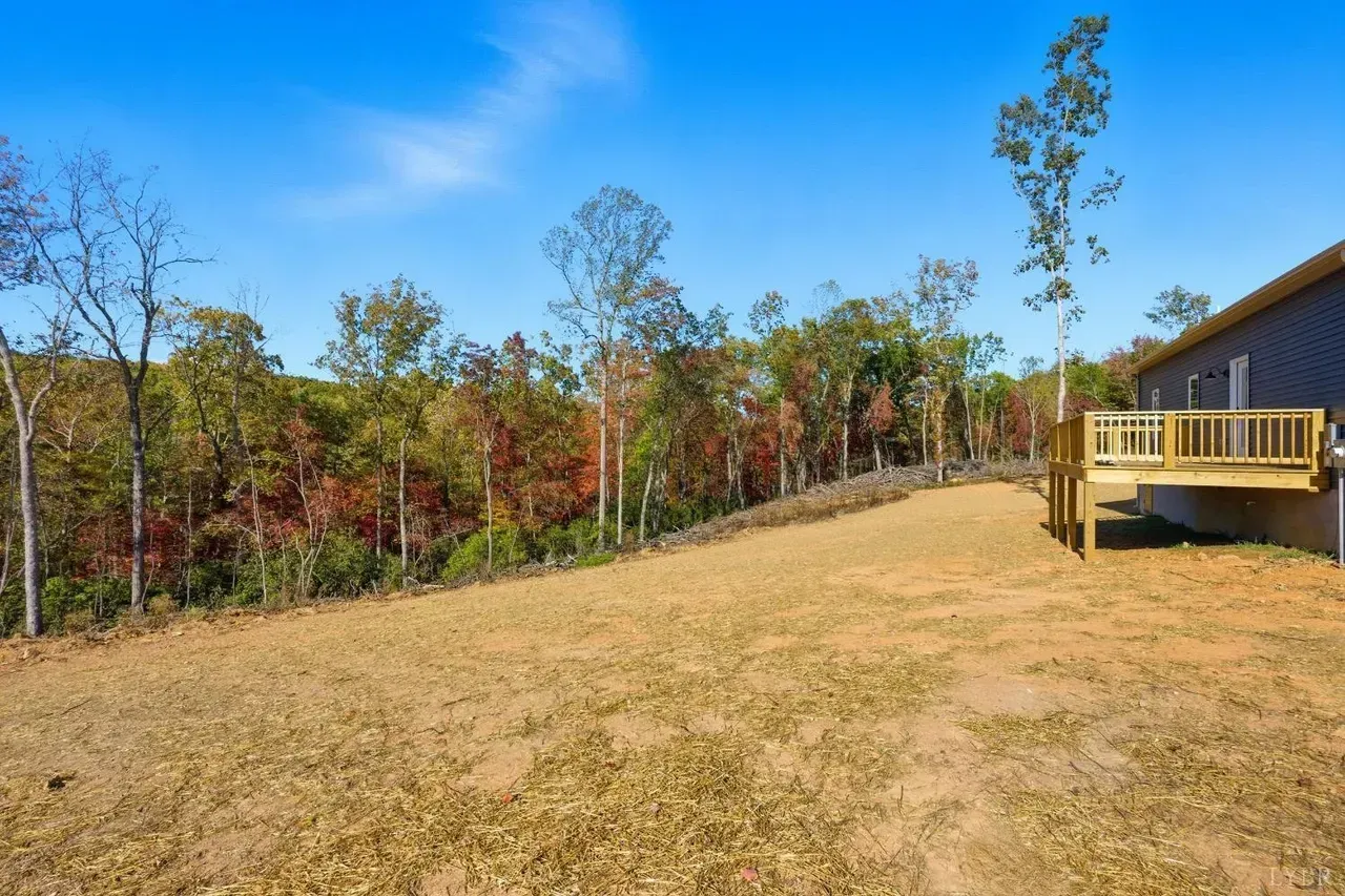 A house deck overlooks a large, sloping grassy backyard bordered by a line of colorful autumn trees under a blue sky.