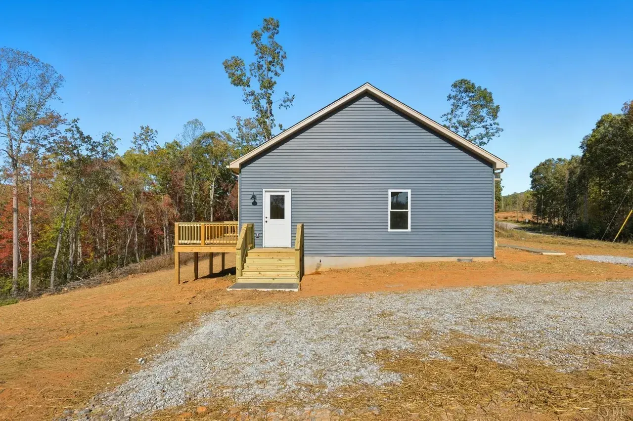 A blue-sided house with a white door and wooden deck stands on a dirt and gravel lot surrounded by woods.