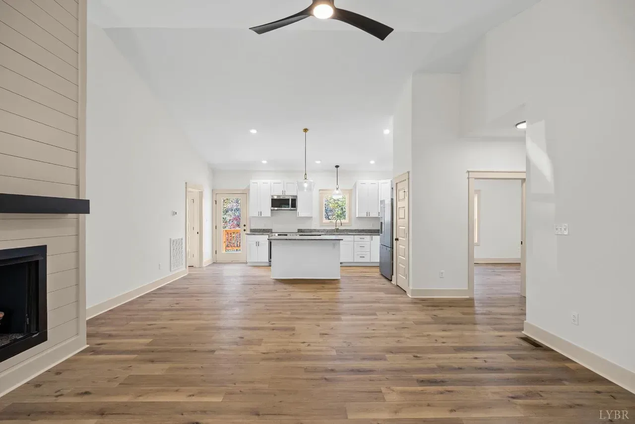A bright, open-concept living area with wood floors, a fireplace on the left, and a white kitchen in the background.
