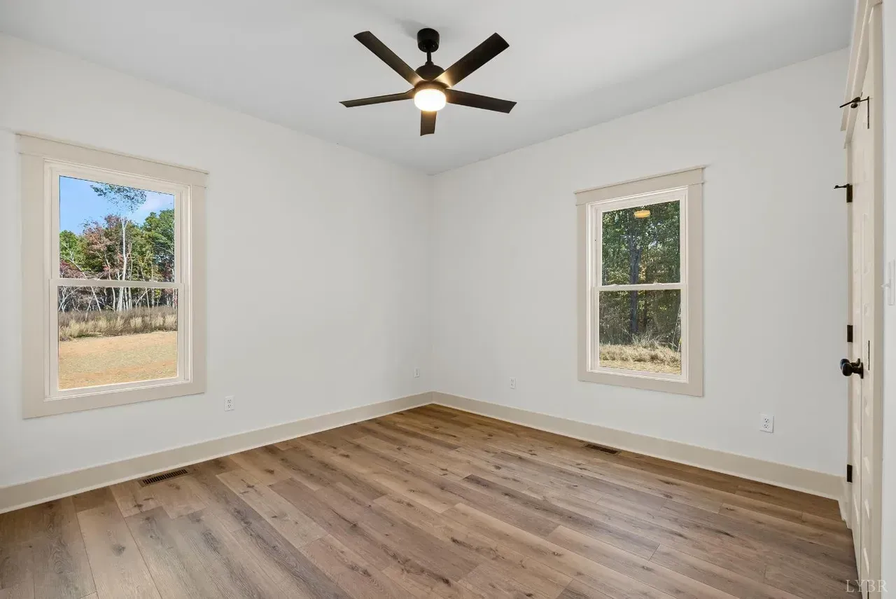 An empty bedroom with white walls, light wood flooring, two windows, and a dark ceiling fan.