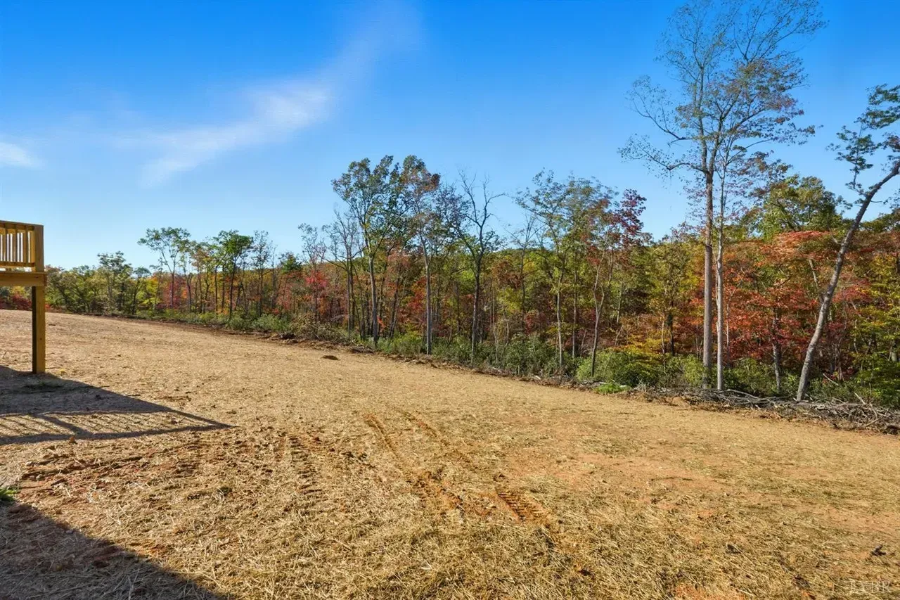 A cleared, dirt-covered yard beside a wooden deck, overlooking a line of trees with autumn foliage under a blue sky.