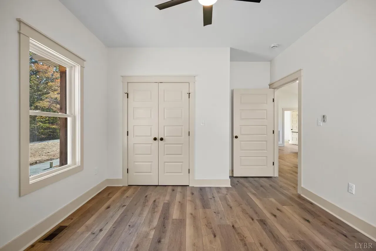A neutral, empty bedroom with white walls, light wood flooring, a window, a double-door closet, and an open doorway.