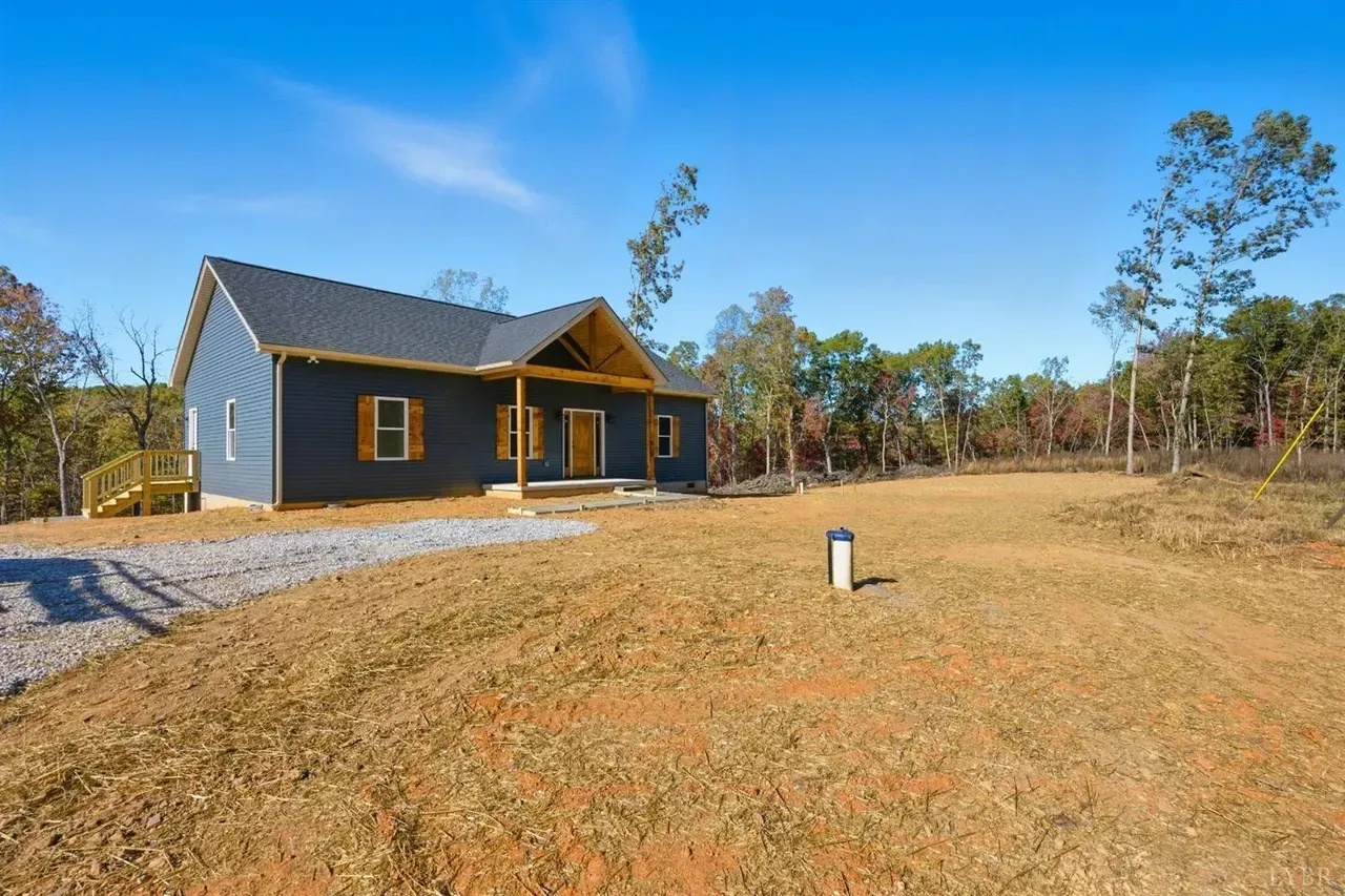 A blue, single-story house with a wooden porch sits on a cleared lot with a gravel driveway under a bright blue sky.