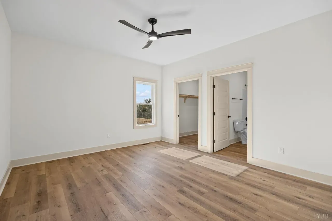 An empty bedroom with white walls, wood-look flooring, a ceiling fan, a window, and two open doorways.