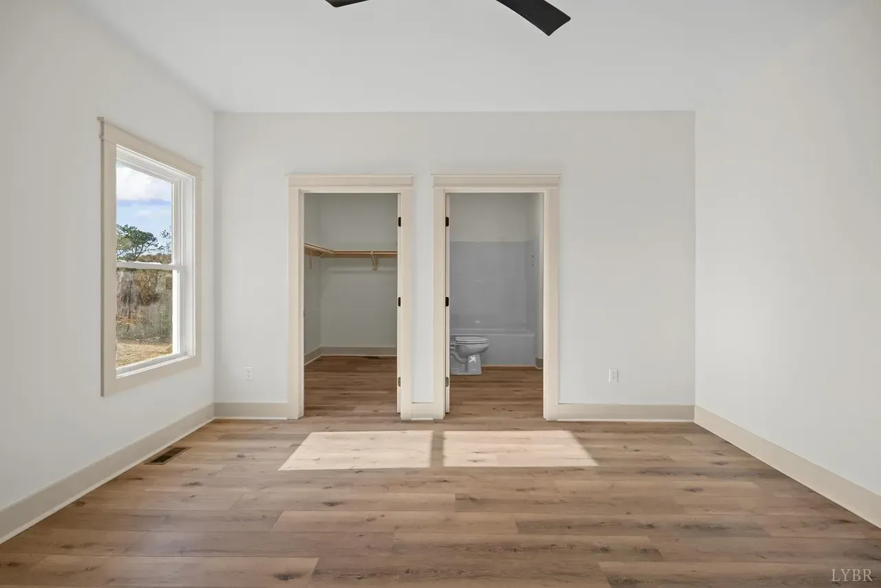 An empty bedroom with light-colored walls, light wood flooring, a window, and two doorways leading to a closet and bathroom.