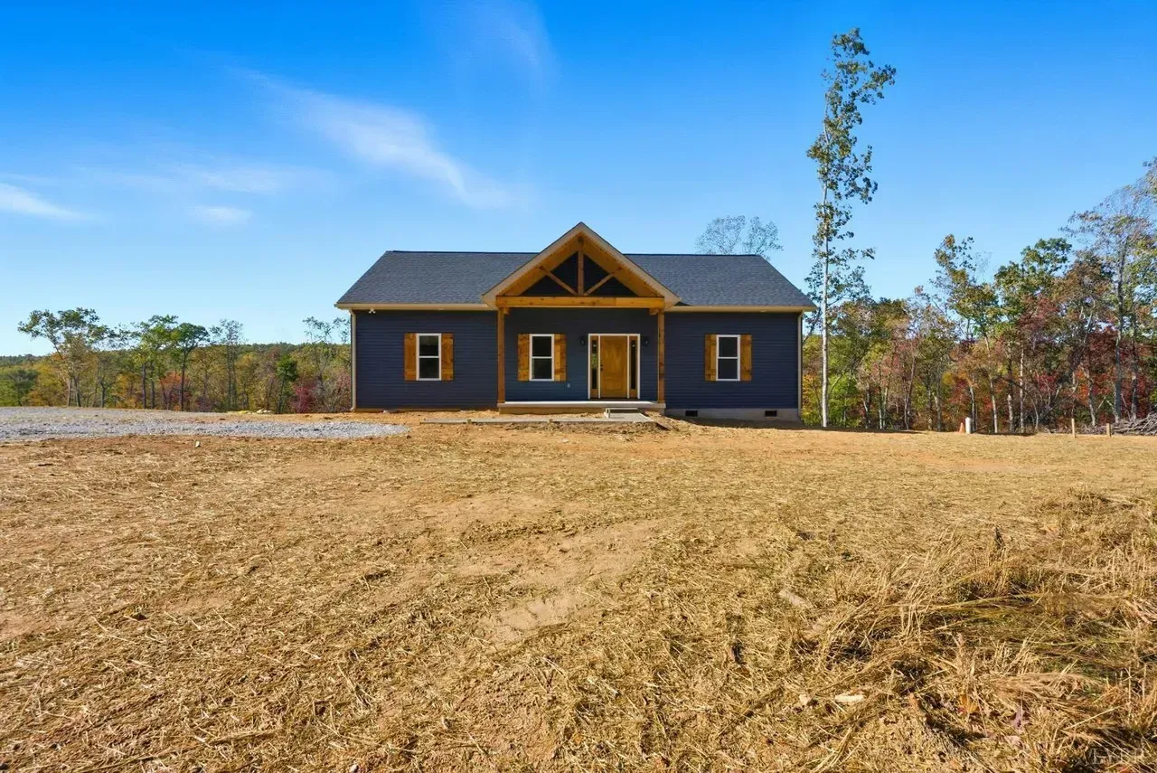 A single-story, dark blue house with a wooden porch gable, situated in a field of dry, sparse grass under a clear blue sky.