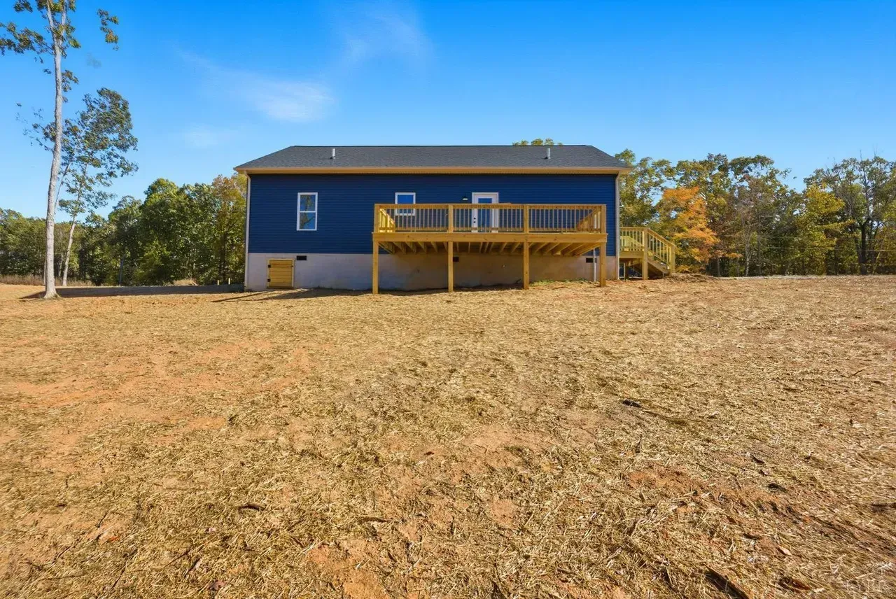 A blue single-story house with a wooden deck stands in a large, dry, open field under a clear blue sky.