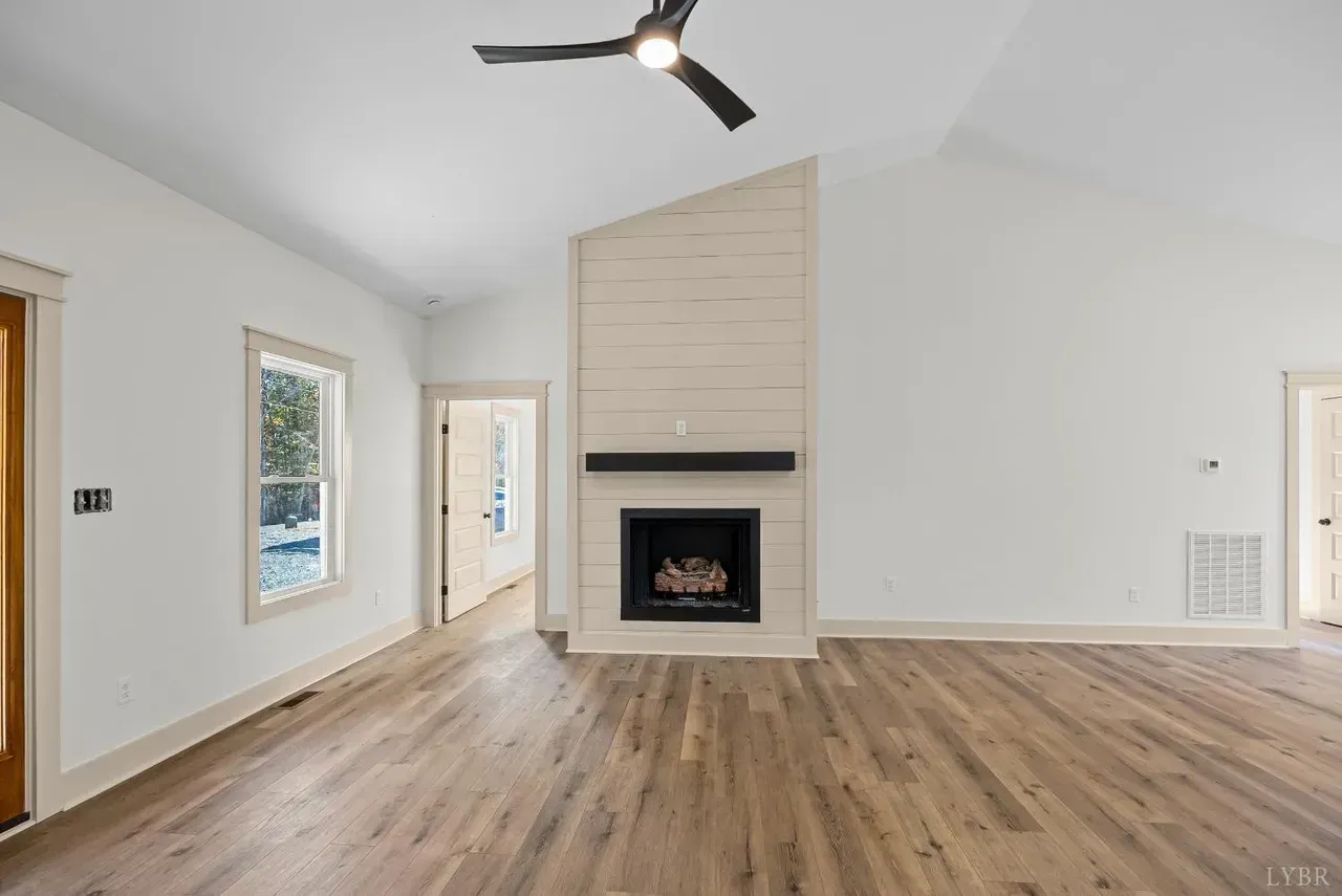 A living room with light wood floors, white walls, a vaulted ceiling with a fan, and a light-toned stone fireplace.