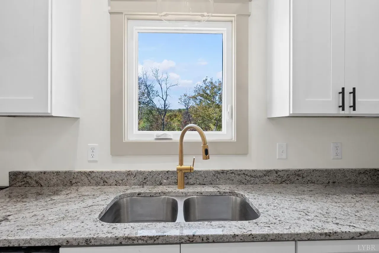 A kitchen sink with a gold faucet in front of a window, flanked by white cabinets and granite countertops.