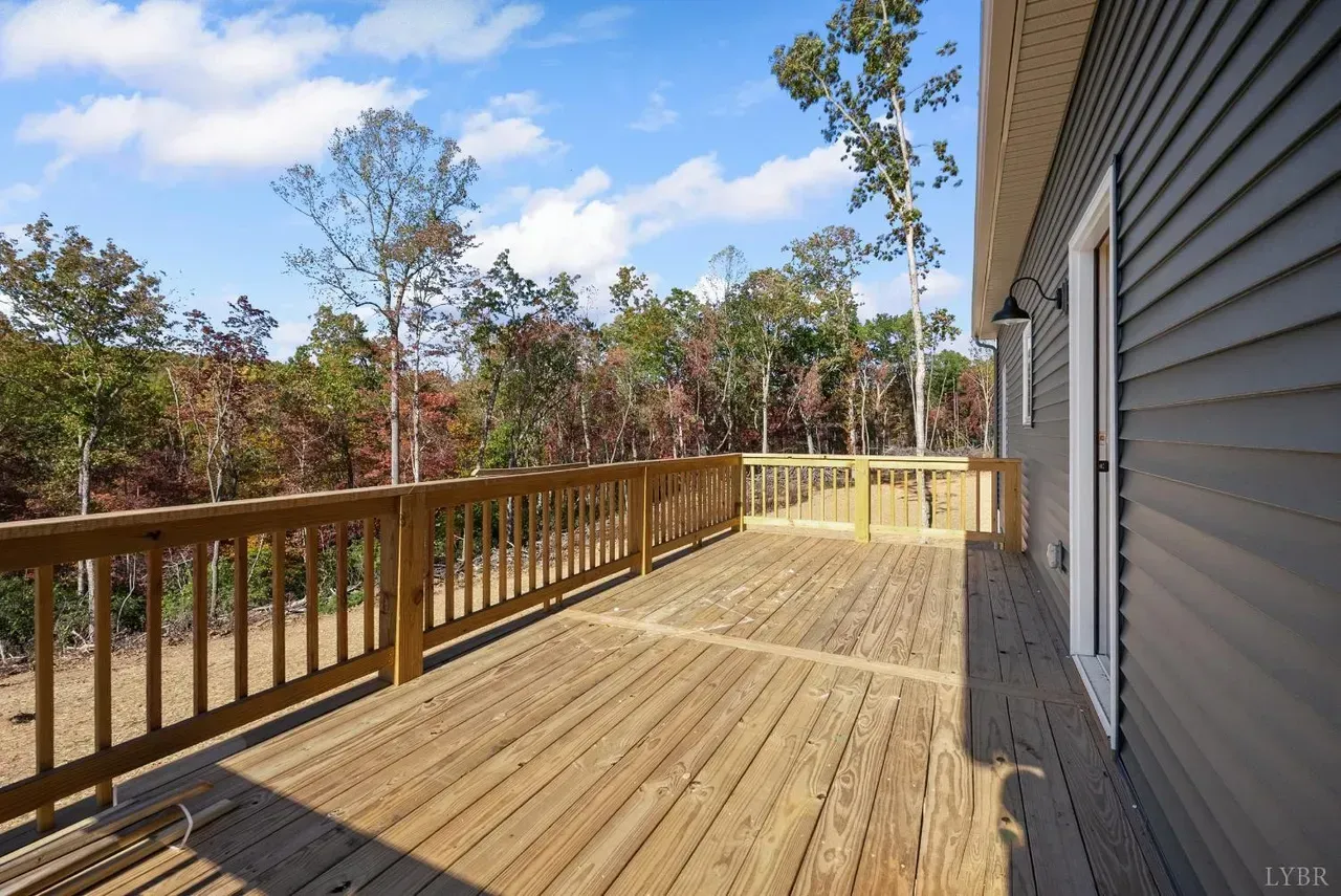 A large wooden deck with railings attached to a grey-sided house, overlooking a wooded area under a blue sky.