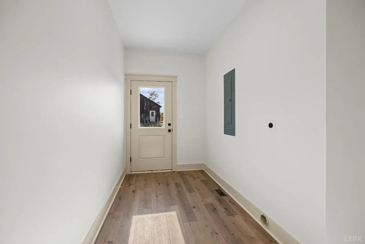 A narrow hallway with light wood-look flooring, white walls, a single door with a window, and a gray electrical panel.
