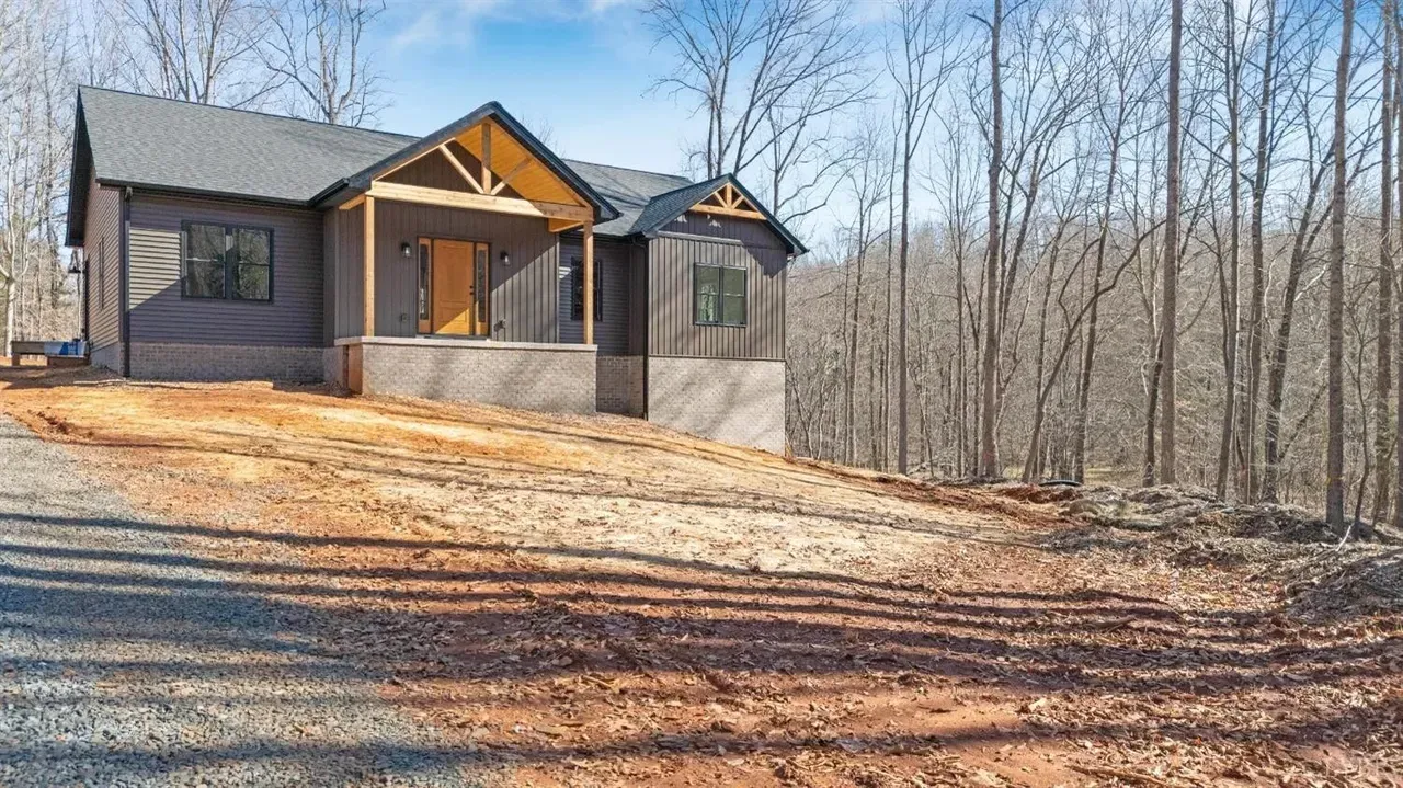 A newly built, dark-sided house with a wooden porch and gabled roof, set on a gravel lot surrounded by a forest.