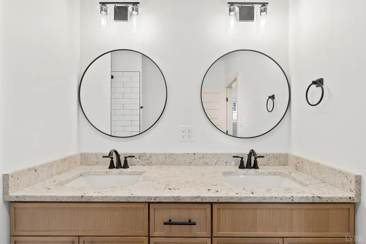 Modern bathroom vanity with light wood cabinets, speckled granite countertop, two sinks, and circular black-rimmed mirrors.