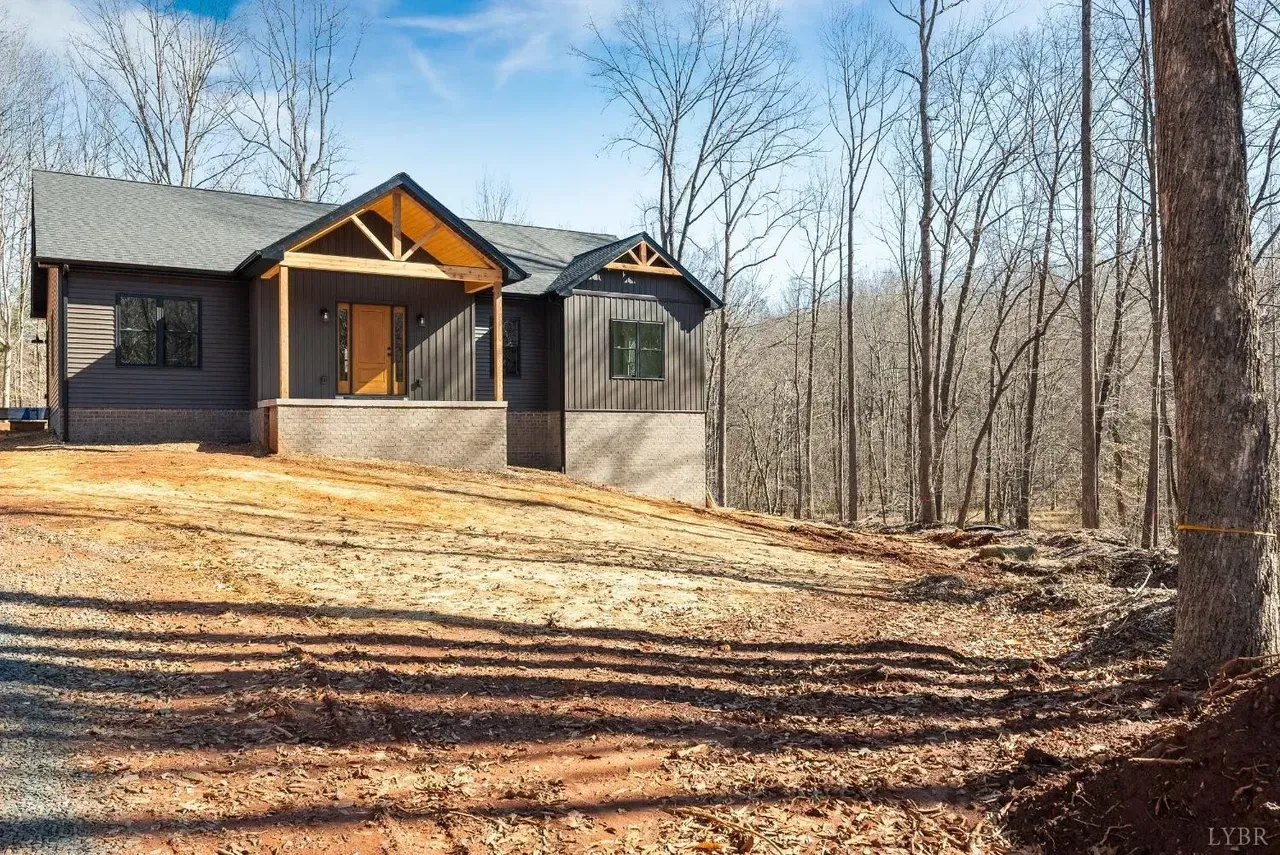 A dark gray house with a wooden porch and brick base sits on a sloped, unlandscaped dirt lot surrounded by trees.