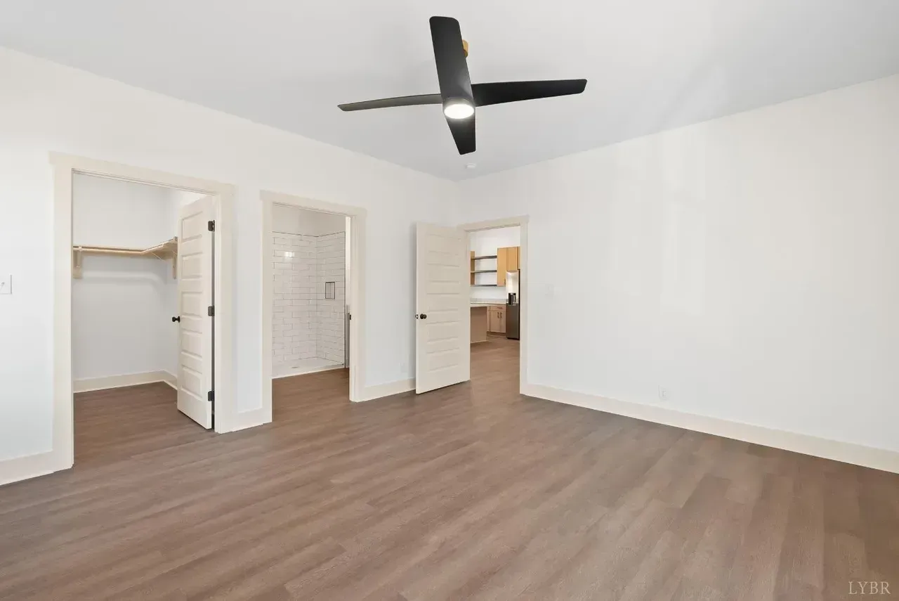 An empty room with white walls, light wood-style flooring, a dark ceiling fan, and three open doorways.