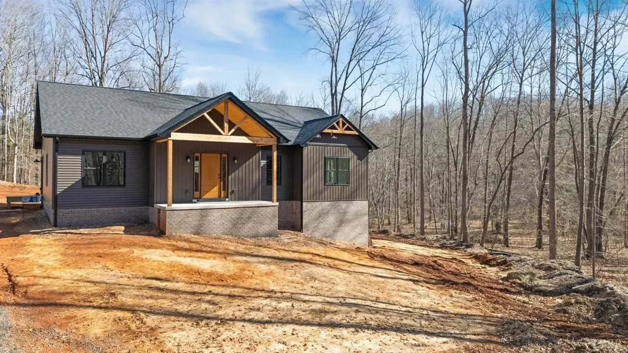 A modern single-story house with dark siding and stone foundation, featuring a timber-framed porch in a wooded setting.