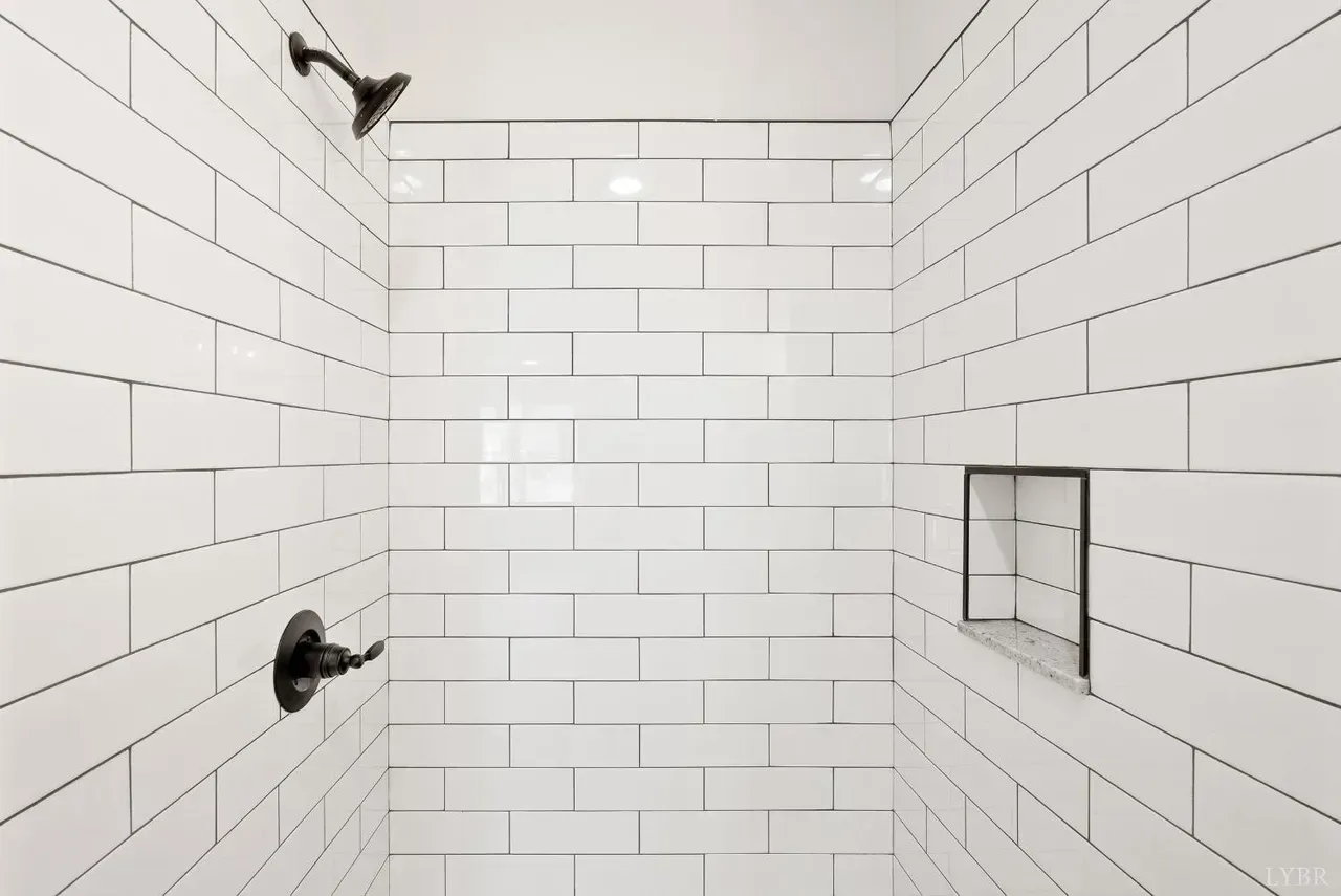 A shower interior featuring white subway tiles, a black showerhead, control handle, and a built-in recessed shelf.